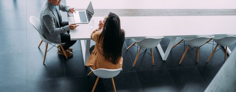 Business partners in meeting. Above view of a two confident business persons sitting on a conference table in the office and sharing ideas. Businessman and businesswoman in meeting using laptop and discussing business strategy. Business coworkers working together in the office.